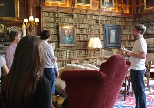 A picture of a tour group inside a grand, classical library at Mostyn Hall, being spoken to by their tour guide, Lord Mostyn.